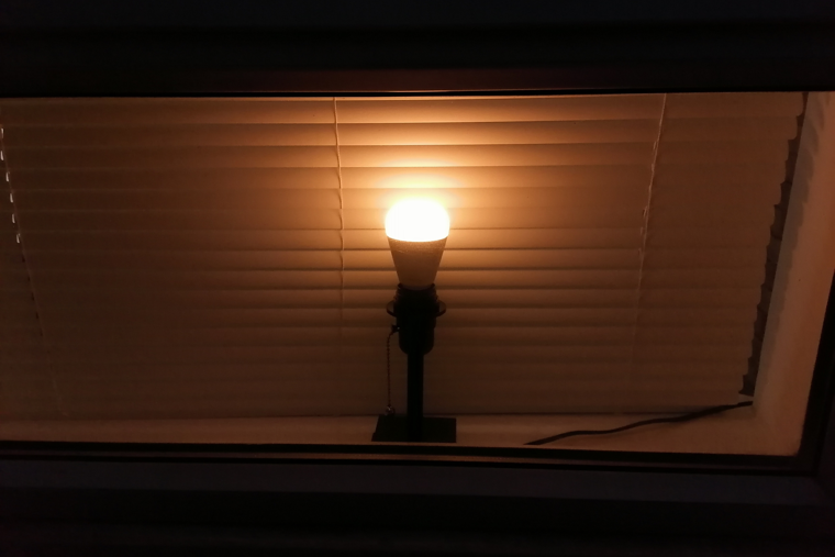 smart bulb lamp glowing warm white in a window in front of closed blinds, seen from outside